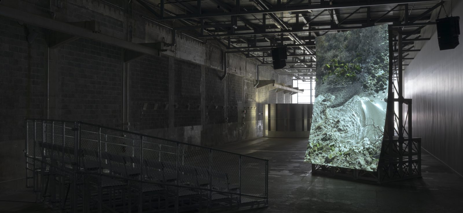 A dimly lit industrial space with metal bleachers faces a large, screen projection. The rocky, forested hillside image enhanced by exposed beams and concrete walls in the stark warehouse atmosphere.