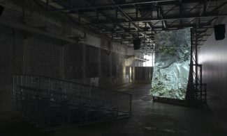 A dimly lit industrial space with metal bleachers faces a large, screen projection. The rocky, forested hillside image enhanced by exposed beams and concrete walls in the stark warehouse atmosphere.