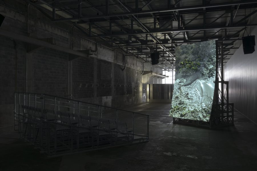 A dimly lit industrial space with metal bleachers faces a large, screen projection. The rocky, forested hillside image enhanced by exposed beams and concrete walls in the stark warehouse atmosphere.