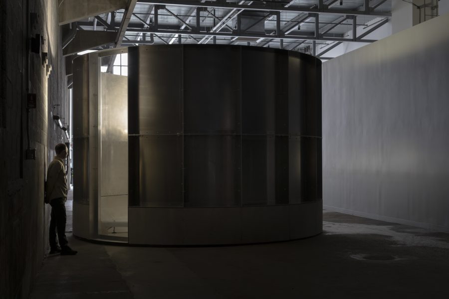 A person stands near a large, cylindrical, metallic structure in a dimly lit industrial space at ICA Boston. Light shines through the glass panels of the structure, reminiscent of Lucy Raven’s "Rounds," casting shadows on the floor.