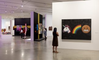 A person stands in a gallery, looking at a colorful painting of a unicorn, rainbow, and emoji on a black background. Other visitors view artworks displayed on black walls in the middle of the space.