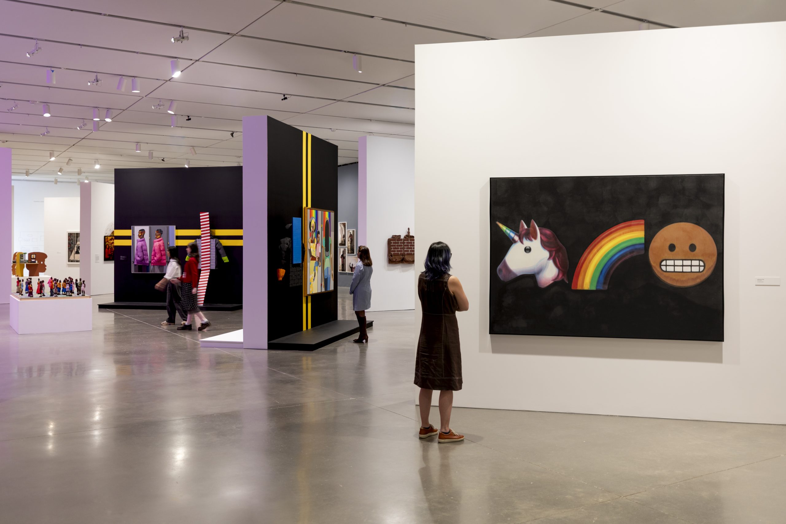 A person stands in a gallery, looking at a colorful painting of a unicorn, rainbow, and emoji on a black background. Other visitors view artworks displayed on black walls in the middle of the space.