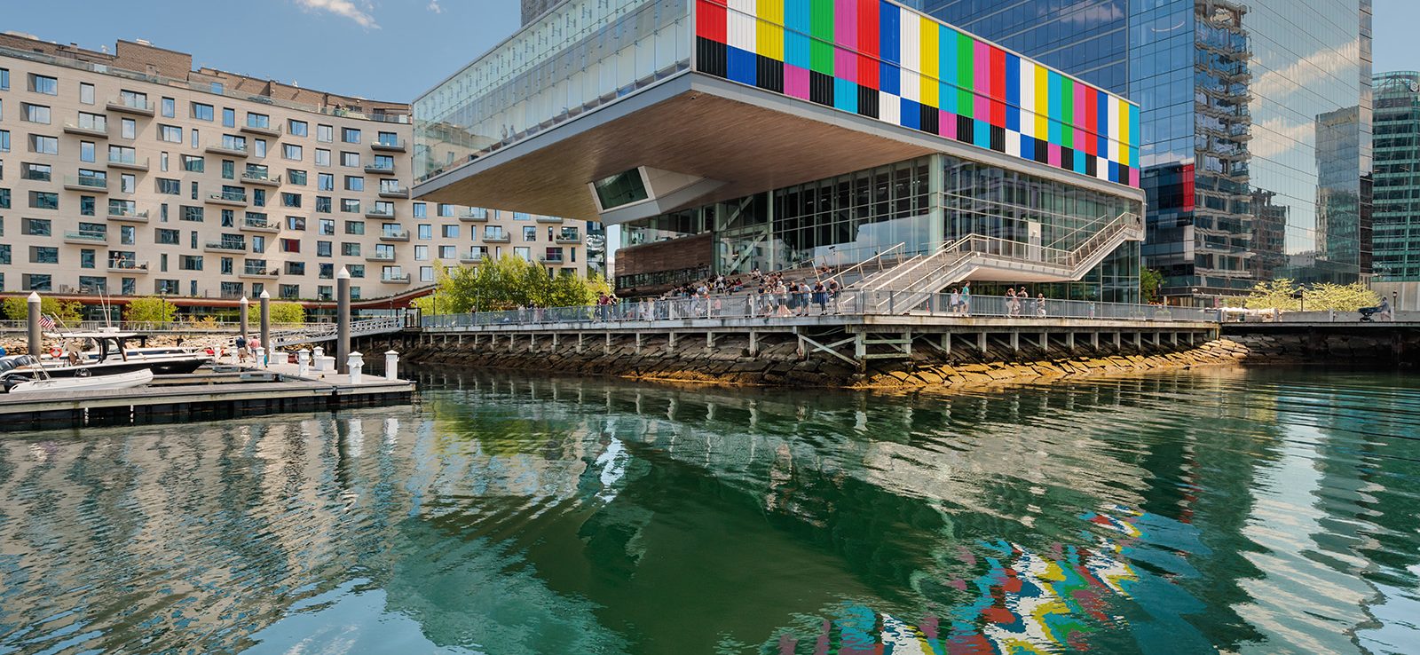 A modern waterfront building with a colorful, striped facade overhangs the harbor at ICA Boston, reflecting in the calm water. People stand on an outdoor terrace enjoying the derrick adams view; boats are docked nearby under a blue sky.