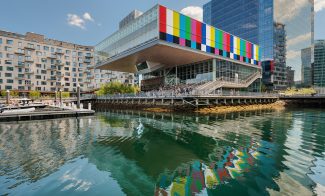 A modern waterfront building with a colorful, striped facade overhangs the harbor at ICA Boston, reflecting in the calm water. People stand on an outdoor terrace enjoying the derrick adams view; boats are docked nearby under a blue sky.