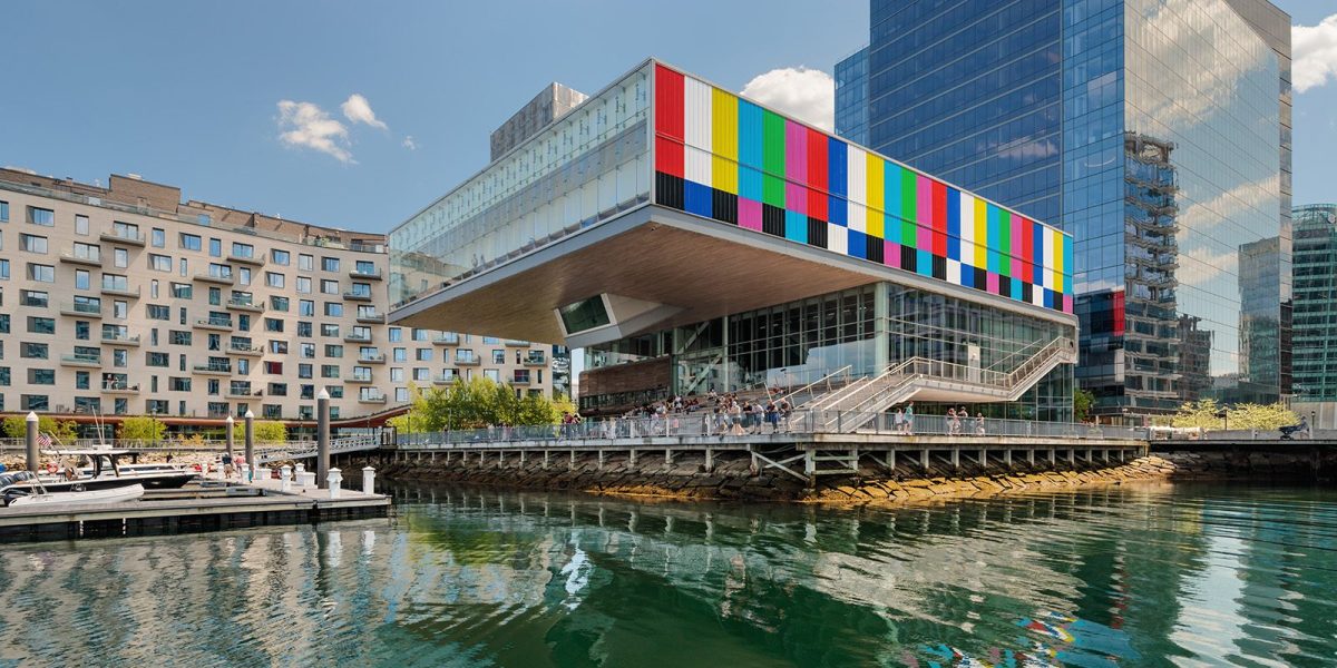 A modern waterfront building with a colorful, striped facade overhangs the harbor at ICA Boston, reflecting in the calm water. People stand on an outdoor terrace enjoying the derrick adams view; boats are docked nearby under a blue sky.