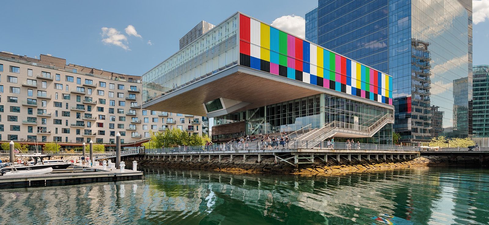 A modern waterfront building with a colorful, striped facade overhangs the harbor at ICA Boston, reflecting in the calm water. People stand on an outdoor terrace enjoying the derrick adams view; boats are docked nearby under a blue sky.