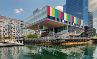 A modern waterfront building with a colorful, striped facade overhangs the harbor at ICA Boston, reflecting in the calm water. People stand on an outdoor terrace enjoying the derrick adams view; boats are docked nearby under a blue sky.
