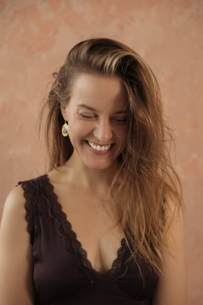A woman with long, light brown hair smiles while looking down. She is wearing a dark lace-trimmed top and a single drop earring, ready to perform at the Summer Stages Dance at ICA. The background is a soft peach color.