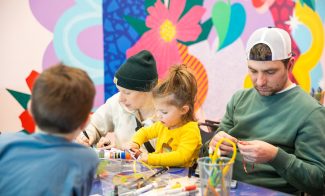 A family sitting at a table doing arts and crafts. An adult and young girl work closely together, while another adult in a cap focuses on a project. Art supplies are scattered across the table; a colorful mural is behind them.