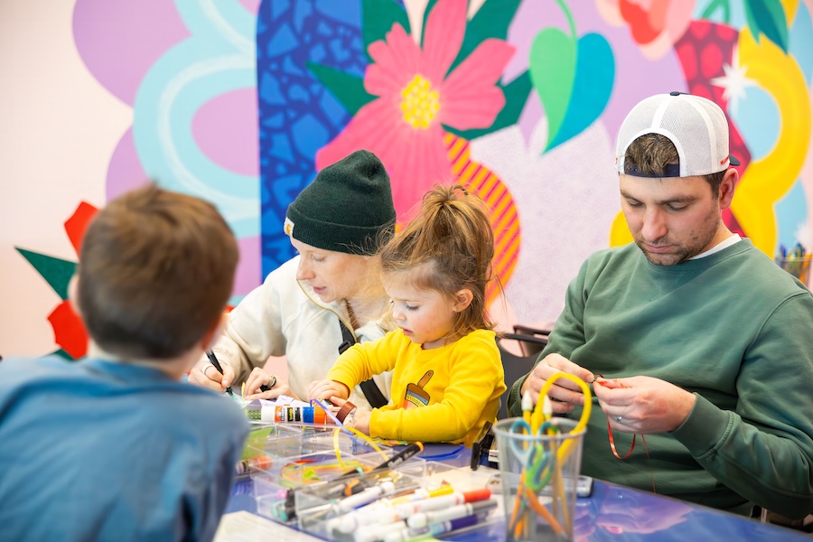 A family sitting at a table doing arts and crafts. An adult and young girl work closely together, while another adult in a cap focuses on a project. Art supplies are scattered across the table; a colorful mural is behind them.