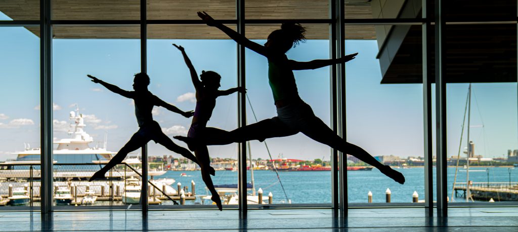 Three dancers perform energetic leaps in a sunlit studio at Summer Stages Dance at ICA, with large windows overlooking a harbor, boats, and the cityscape beyond.