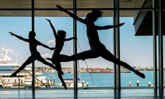 Three dancers perform energetic leaps in a sunlit studio at Summer Stages Dance at ICA, with large windows overlooking a harbor, boats, and the cityscape beyond.
