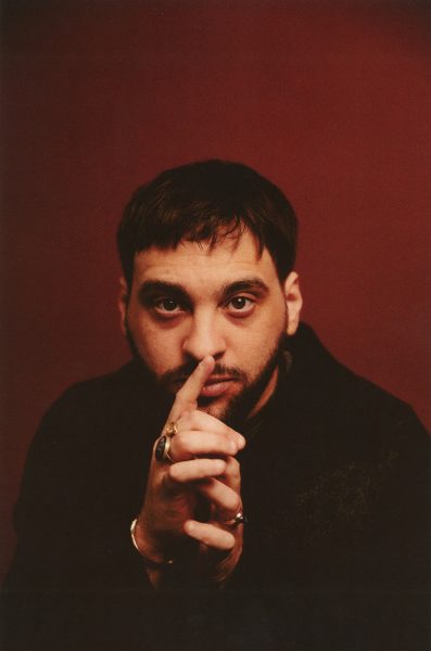 A person with short dark hair and a beard sits against a deep red background, gazing at the camera with their finger resting vertically against their lips—evoking the intrigue of Summer Stages Dance at ICA. They wear rings, bracelets, and a dark top.