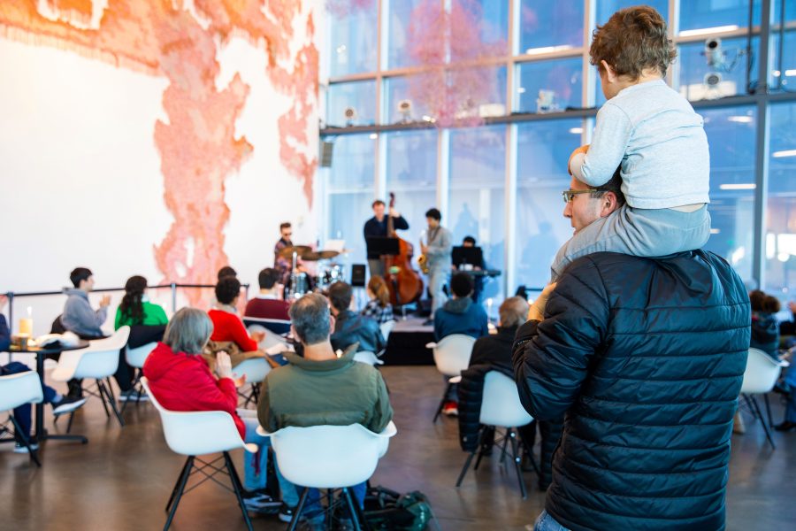 A family-friendly concert features musicians playing on a small stage in a bright room. Seated attendees watch, while a man stands with a child on his shoulders. Large windows and abstract wall art are visible in the background.