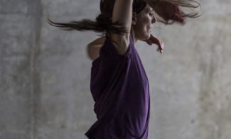 A person with long hair wearing a purple shirt and dark pants is mid-motion, dancing energetically with arms raised against a gray concrete background, capturing the spirit of Summer Stages Dance at ICA.
