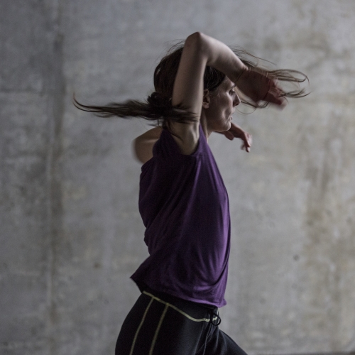 A person with long hair wearing a purple shirt and dark pants is mid-motion, dancing energetically with arms raised against a gray concrete background, capturing the spirit of Summer Stages Dance at ICA.