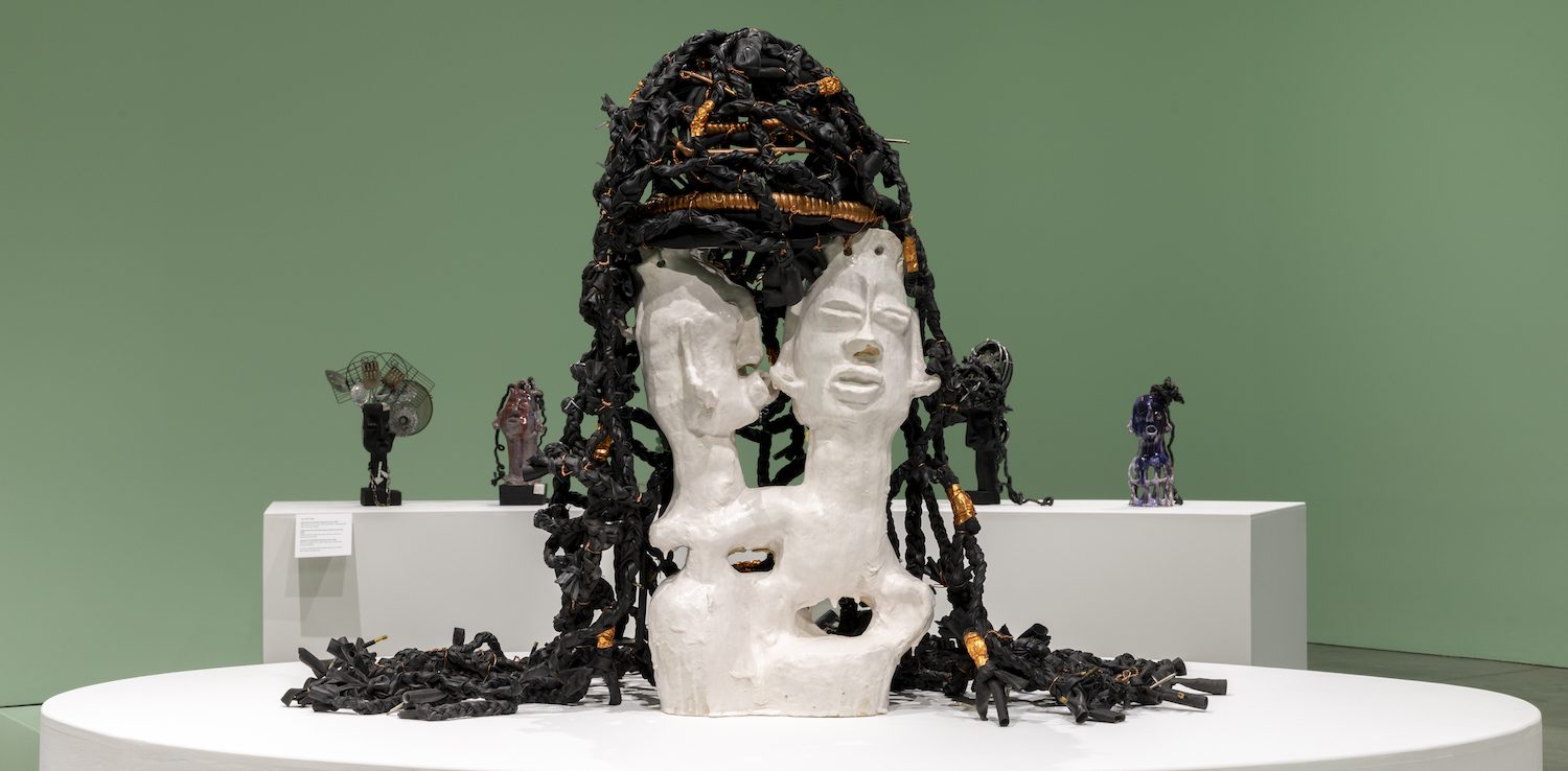 A white sculpted bust with two faces and black and orange textured material resembling hair—reminiscent of Leilah Babirye’s distinctive style—sits on a round white pedestal in a green-walled gallery at ICA Boston, with other abstract sculptures in the background.