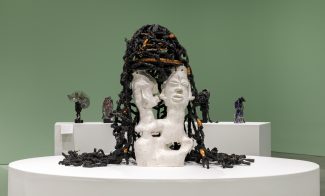 A white sculpted bust with two faces and black and orange textured material resembling hair—reminiscent of Leilah Babirye’s distinctive style—sits on a round white pedestal in a green-walled gallery at ICA Boston, with other abstract sculptures in the background.
