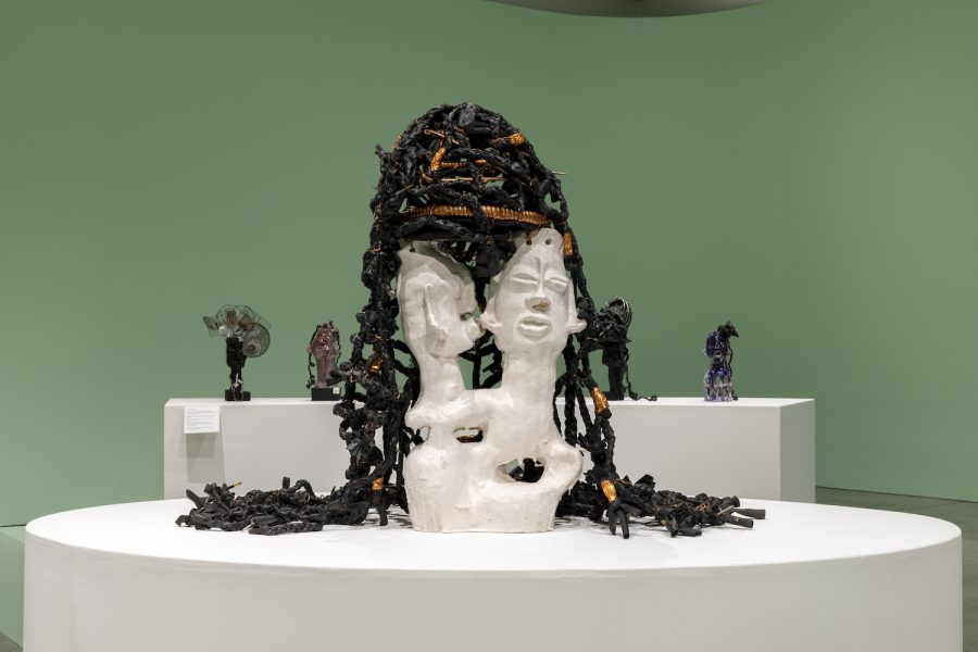 A white sculpted bust with two faces and black and orange textured material resembling hair—reminiscent of Leilah Babirye’s distinctive style—sits on a round white pedestal in a green-walled gallery at ICA Boston, with other abstract sculptures in the background.