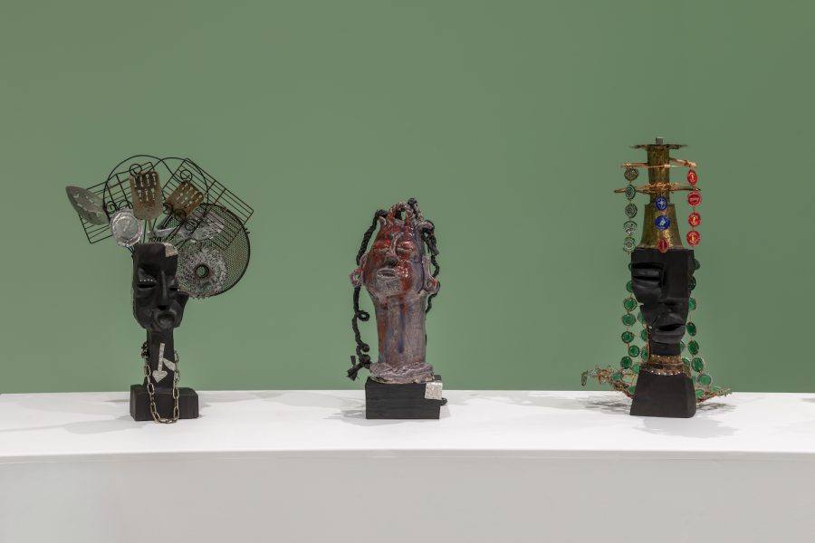 Three abstract sculptures resembling heads, crafted from various materials like metal, wire, beads, and plastic, are displayed on a white pedestal against a solid green background—evoking the innovative spirit of Leilah Babirye’s works as seen at ICA Boston.