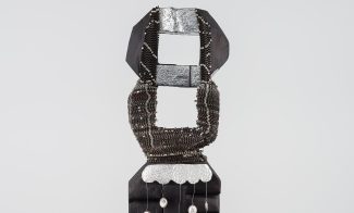 A tall, abstract black sculpture with geometric shapes, metallic accents, beaded chains, and stirrup-like objects hanging from each side stands against a white background—reminiscent of works by Leilah Babirye at ICA Boston.