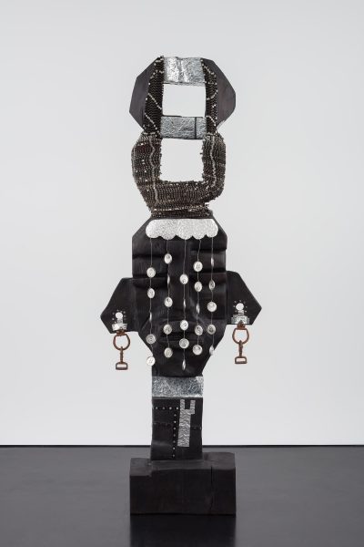 A tall, abstract black sculpture with geometric shapes, metallic accents, beaded chains, and stirrup-like objects hanging from each side stands against a white background—reminiscent of works by Leilah Babirye at ICA Boston.