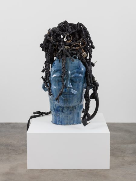 A blue sculpted head with closed eyes sits on a white pedestal. The textured hair, crafted from black braided materials and objects, evokes leilah babirye’s style—making this piece a striking highlight at ICA Boston.