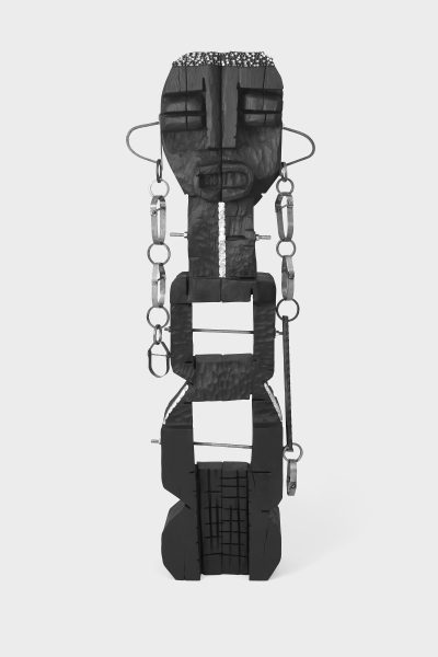 A tall, abstract black sculpture resembling a human figure with geometric shapes, textured surfaces, and various metal rings and wires attached to its sides, stands against a plain white background—reminiscent of Leilah Babirye’s works at ICA Boston.
