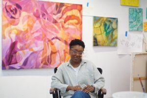 A person wearing glasses and a light shirt sits in front of a wall covered with colorful abstract paintings and sketches, capturing the creative spirit of artmaking after dark at ICA Boston.