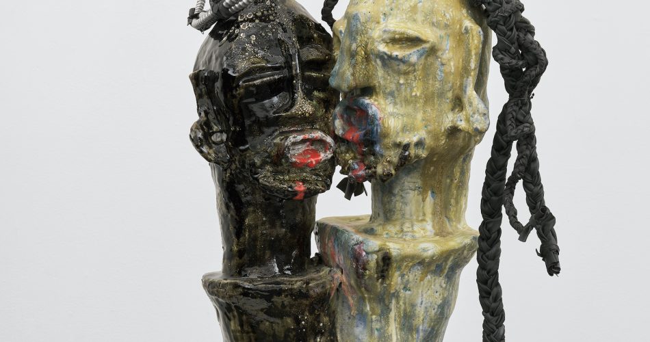 Two abstract, faceless busts—one dark, one yellow—lean together on a pedestal. The dark figure has coiled metal on its head; the yellow figure has twisted black cords as hair. Both have textured, rough surfaces.