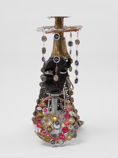 A mixed-media sculpture inspired by Leilah Babirye, recently exhibited at ICA Boston, featuring a dark mask, bottle caps, metal, and wire arranged on a stand, with colorful accents and hanging bottle cap chains against a plain white background.