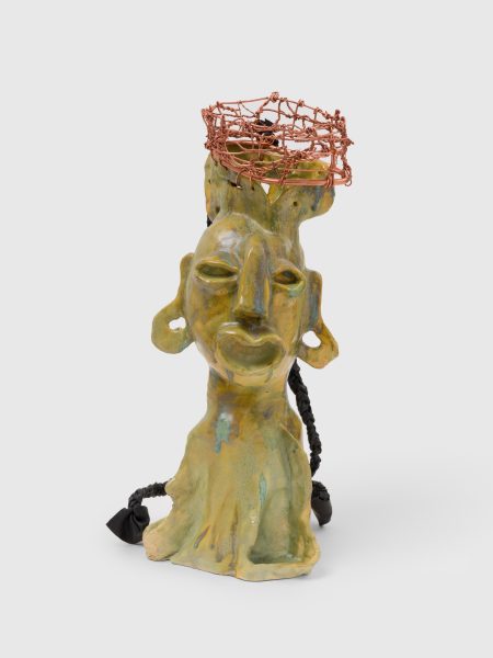 A yellow-green abstract ceramic sculpture of a face with large ears, a wide nose, braided black cords, and a copper wire basket on top of the head, reminiscent of Leilah Babirye’s distinctive style, set against a plain white background. This striking piece evokes the innovative contemporary art found at ICA Boston.
