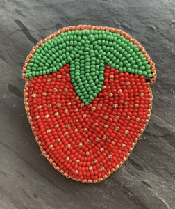 A beaded brooch shaped like a strawberry, with red and green beads, displayed on a dark gray surface—perfect for adding a touch of artmaking after dark style reminiscent of ICA Boston.