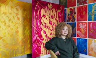 A woman with curly hair sits on a chair in front of colorful abstract paintings with bold calligraphic designs in yellow, pink, and a mosaic of red, blue, and orange panels.