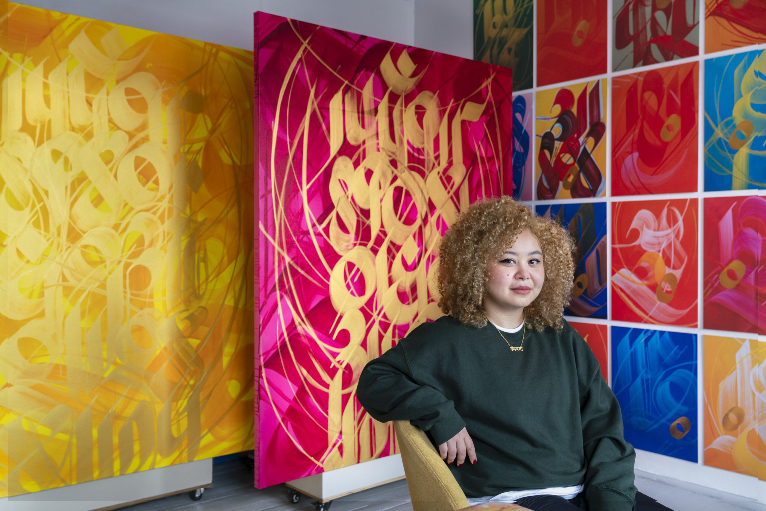 A woman with curly hair sits on a chair in front of colorful abstract paintings with bold calligraphic designs in yellow, pink, and a mosaic of red, blue, and orange panels.