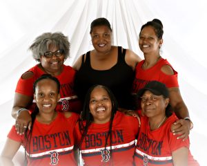 Six women pose together, five in matching red Boston T-shirts and one in a black tank top, all smiling in front of a white draped background at an artmaking after dark event at ICA Boston.