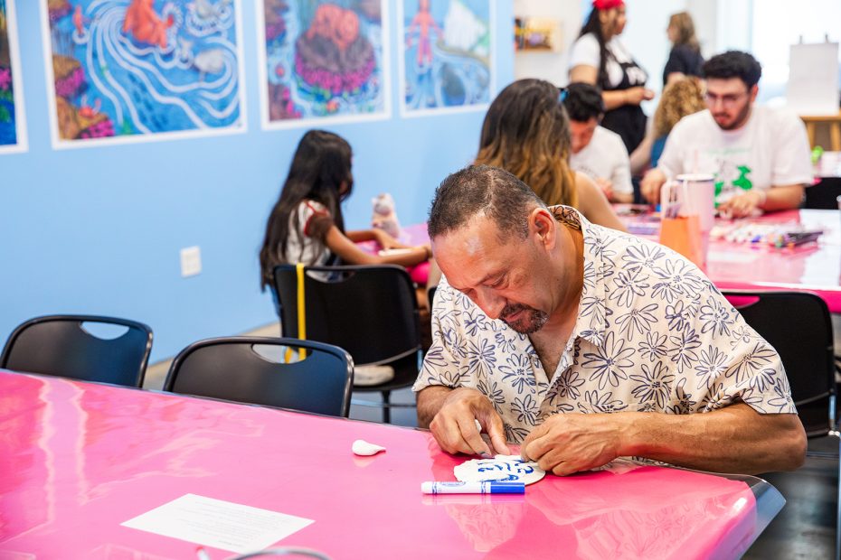 A man concentrates on coloring a craft at a table covered with a pink tablecloth, while others, including children, engage in creative activities in a bright, art-filled room.
