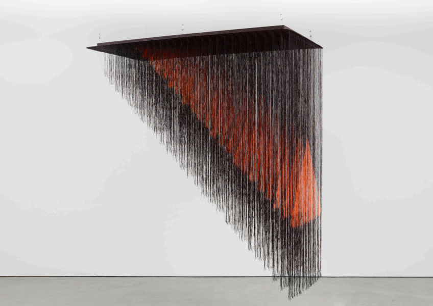 A geometric installation of hanging black strings forms a suspended triangular shape from the ceiling, with an orange gradient pattern visible among the strands against a white wall.