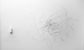 A small white box attached to a wall releases a pen that draws a chaotic, looping pattern of black lines across the otherwise blank, white surface.
