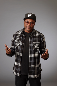 A man wearing sunglasses, a black-and-white plaid shirt over a black t-shirt, black pants, and a black baseball cap with a white P logo stands against a gray background, dressed as if ready for the ICA Gala 2025, with his hands slightly raised.