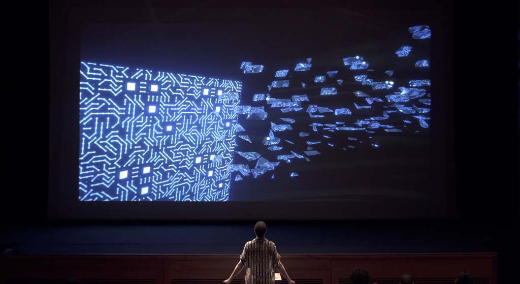 A person stands facing a large digital screen displaying a glowing circuit board pattern on the left and scattered floating digital shards on the right, resembling data emerging from the circuitry.