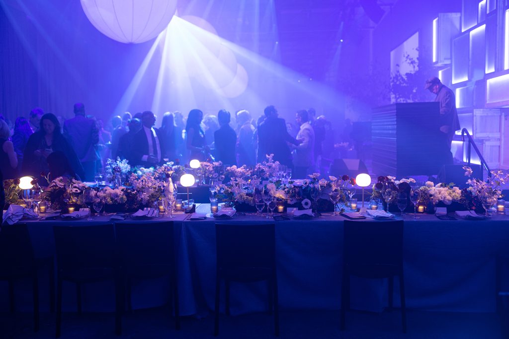 A dimly lit event space at the ICA Gala, where people dance and mingle in blue and purple lighting. In the foreground, a long table is adorned with flowers, candles, and glowing orbs.