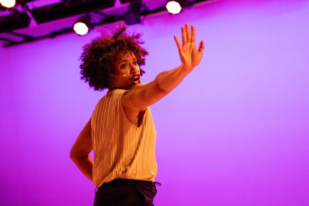 A woman with curly hair, wearing a sleeveless top and headset microphone, gestures with an outstretched hand while performing on stage with purple lighting.