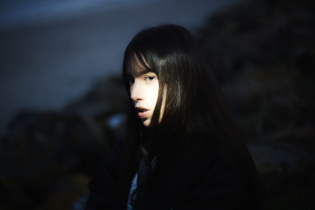 A woman with long dark hair, her face partially illuminated by a beam of light, with a soft-focus background of rocks and dim surroundings.