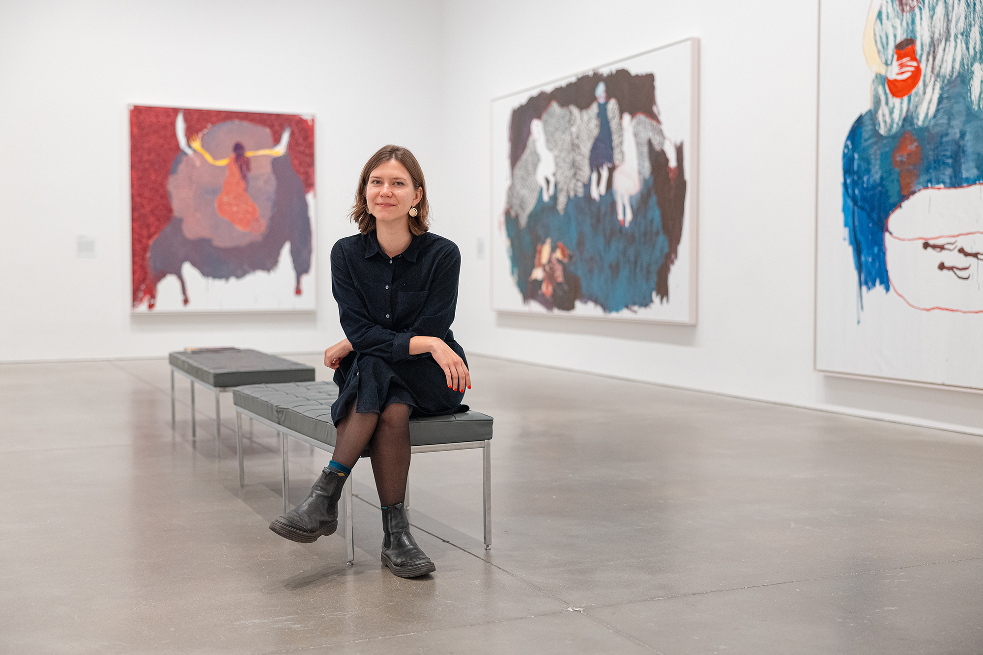 A woman in a black dress and boots sits on a bench in ICA/Boston, surrounded by large colorful paintings on white walls