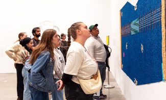 A group of people stands closely together in a gallery, intently observing a large, textured blue artwork displayed on a white wall. Some lean forward to examine the details during the tour.