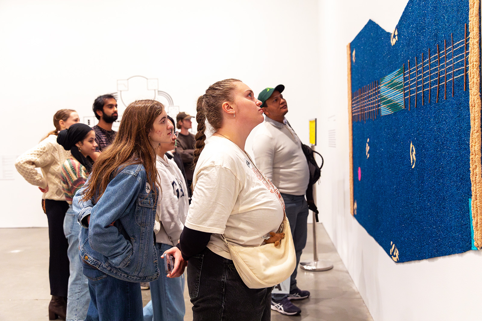 A group of people stands closely together in a gallery, intently observing a large, textured blue artwork displayed on a white wall. Some lean forward to examine the details during the tour.