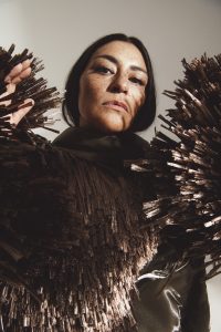 A person with long dark hair and freckles wears a dramatic, metallic, spike-embellished outfit.