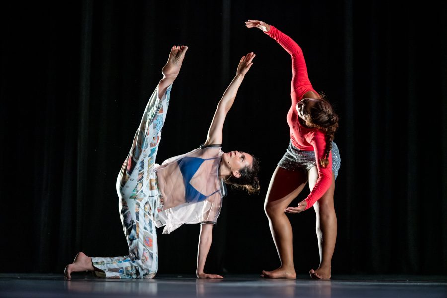 Two dancers from Rashaun Mitchell + Silas Riener: Open Machine strike expressive poses on stage; one balances on one hand with a high leg extension, while the other arches gracefully towards her against a black background.