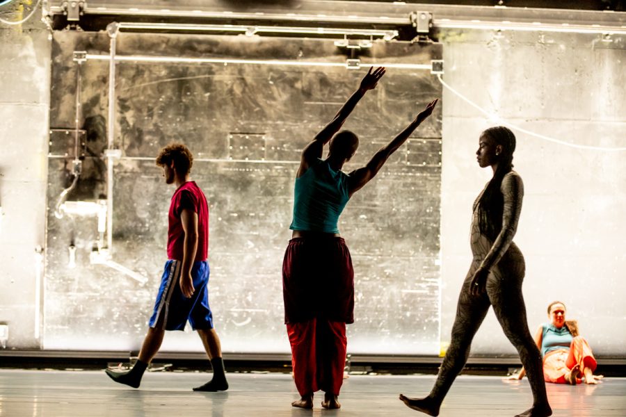 Four people on a stage evoke dynamic motion in the dramatic, artistic atmosphere of Rashaun Mitchell + Silas Riener: Open Machine, with one standing arms raised and another walking past with long braids as others move in the background.